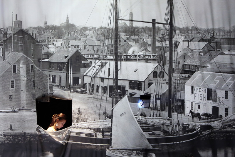 Joclyne Nunes, programs manager, is seen operating the projector and slideshow from behind an historical photo of New Bedford's waterfront, at the back of the Whaling Museum auditorium during the sixth annual Gateway Cities Innovation Awards. PHOTO PETER PEREIRA