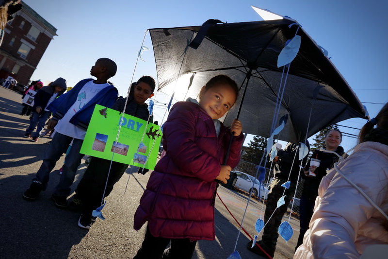 Smiling to her parents who are looking on, Sofia DaCosta, and fellow first graders represent antonyms (wet/dry), as Swift School students and staff participate in the annual Vocabulary Figurative Language Parade, holding signs and dressed in costume, showcasing their vocabulary skills in a parade around the New Bedford, MA elementary school.   PHOTO PETER PEREIRA