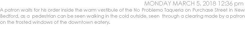 A patron waits for his order inside the warm vestibule of the No Problemo Taqueria on Purchase Street in New Bedford, as a  pedestrian can be seen walking in the cold outside, seen  through a clearing made by a patron on the frosted windows of the downtown eatery.