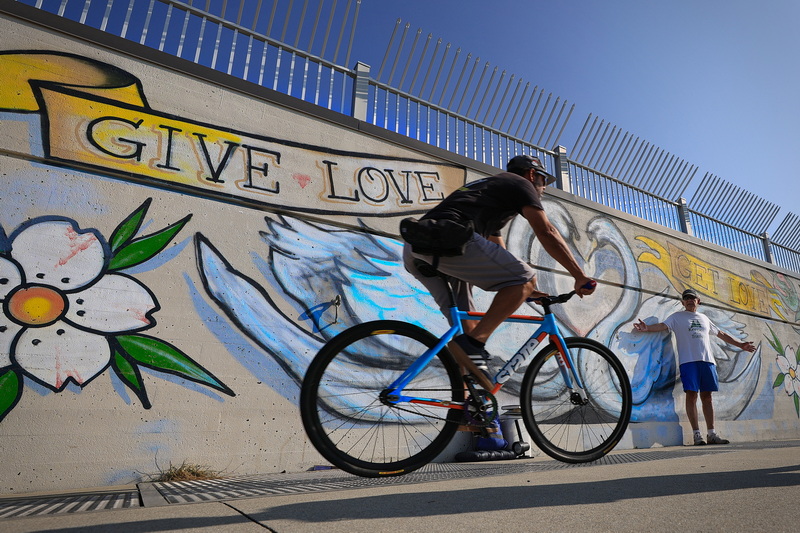 A man gives way to a man riding a bicycle on the Blue Lane walk atop the hurricane barrier overlooking Clarke's Cove in New Bedford, MA behind them a colorful mural.  PHOTO PETER PEREIRA