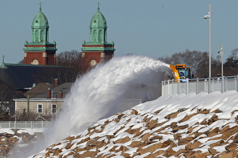 In the distance, the steeples of Mount Carmel church are visible while a New Bedford DPI employee operates a snowblower, sending snow flying into the air as he clears the Clark's Cove Walk atop the hurricane barrier in the south end of New Bedford, MA. PHOTO PETER PEREIRA
