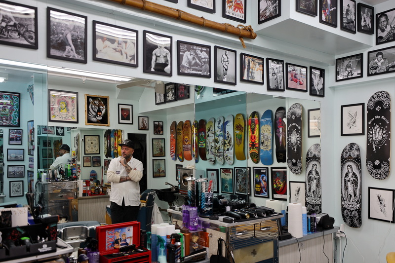 Danny Baptista, owner, is pictured among photographs and skateboards, his reflection visible in the mirrors of his Stepping Razor Barber Shop in downtown New Bedford as he pauses between clients. PHOTO PETER PEREIRA