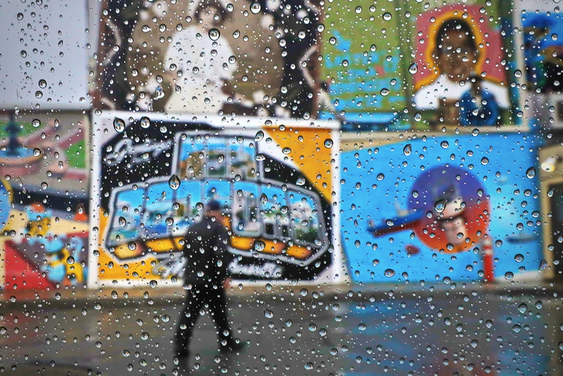 The colorful murals are seen in the background as a man walks across the Bank of America parking lot in downtown New Bedford, MA on a rainy day as seen through the rain drops on a car window. PHOTO PETER PEREIRA