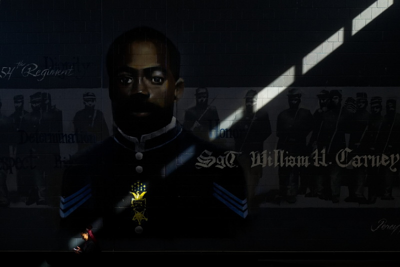 A beam of light coming from the windows above shines on a teacher as she walks past a mural of Carney Academy Elementary School's namesake, SGT William H. Carney, the first African American to receive the Medal of Honor. William Harvard Carney was awarded the Medal of Honor in 1900 for his gallantry during the Civil War at the Battle of Fort Wagner in 1863 and is buried at the Oak Grove Cemetery in New Bedford, MA. PHOTO PETER PEREIRA